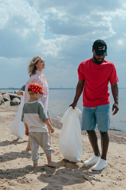 A man and two children on a sandy beach during the daytime, engaging in rubbish collection activities. The man, dressed in a red t-shirt, light blue denim shorts, and a black cap, is holding a white plastic bag and appears to be picking up litter from the ground. A young girl with curly blonde hair, wearing a light pink jacket, stands nearby, watching the activity. A young boy in a camouflage-patterned cap, a light grey t-shirt, and beige pants is also present, holding a white plastic bag open as if preparing to collect trash. In the background, the scene shows a calm body of water extending to the horizon under a partly cloudy sky, with rocks lining the shoreline. The setting indicates an outdoor environment suitable for private waste handling or environmental cleanup, which relates closely to rubbish removal services such as those offered by Rubbish Removal Swiss Cottage.