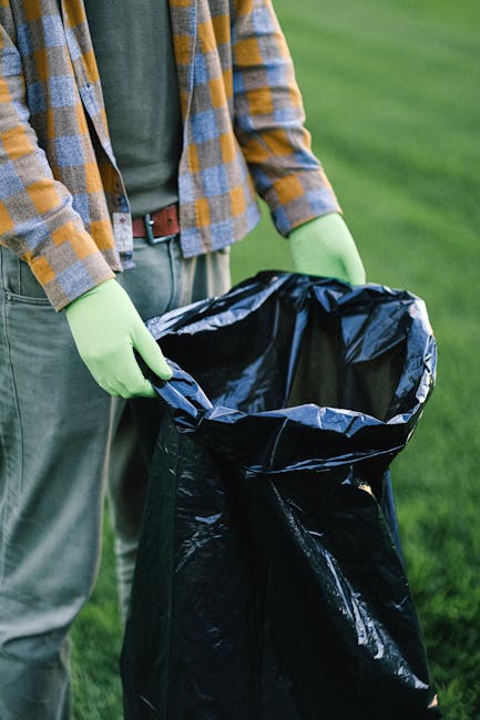A person standing outdoors on a grassy area, wearing a plaid shirt with shades of yellow, blue, and gray, and green gloves, holding open a large black rubbish bag made of plastic. The bag appears to be partially filled, with the top edge folded outward, revealing the dark interior. The person's lower body is visible, dressed in gray trousers with a brown belt. The background shows a lush, green lawn, indicating a garden or park setting. The scene suggests a waste disposal activity, such as rubbish collection or garden clearance, supported by the presence of the rubbish bag and gloves suitable for handling waste, which aligns with independent waste handling or alternative rubbish removal services.