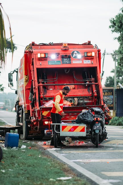 A worker in a high-visibility orange vest and dark trousers is standing next to the rear of a large red waste collection truck, which features various safety and identification stickers, including a serial number and hazard symbols. The worker is operating or inspecting the waste collection mechanism at the back of the truck, which has an open hatch revealing a metal interior designed for compacting rubbish. The scene is set on a paved roadside with a designated waste collection area, evident from the curb and parking markings. Nearby, a black motorcycle with a storage bag attached is parked close to the truck. Scattered debris and a small blue container are visible on the ground, suggesting recent rubbish collection activity. The area has some greenery, with trees and bushes in the background, and an overcast sky, providing diffuse natural light. The environment and equipment indicate an on-site waste collection or rubbish removal process, consistent with private rubbish handling services such as those provided by Rubbish Removal Swiss Cottage.