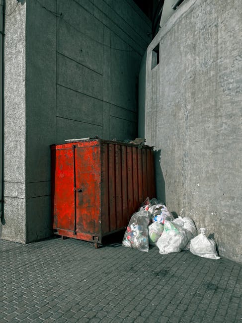Three large black plastic bags filled with waste material are placed on the pavement near the curb, leaning against a black metal fence with vertical bars. The bags appear to contain household or shop rubbish and are loosely tied at the top, with some parts of the plastic wrinkled and reflective under diffuse natural light. Behind the fence, there is foliage from leafy trees or shrubs, adding a textured green backdrop to the scene. The pavement is smooth with a slightly raised edge adjacent to a dark asphalt road. The setting appears to be an outdoor urban or suburban area, possibly in front of shops or cafes, consistent with private waste collection or rubbish removal services. The overall atmosphere is quiet and tidy, with no human activity visible, highlighting a typical instance of discarded rubbish awaiting collection or disposal through independent waste management such as that provided by Rubbish Removal Swiss Cottage.