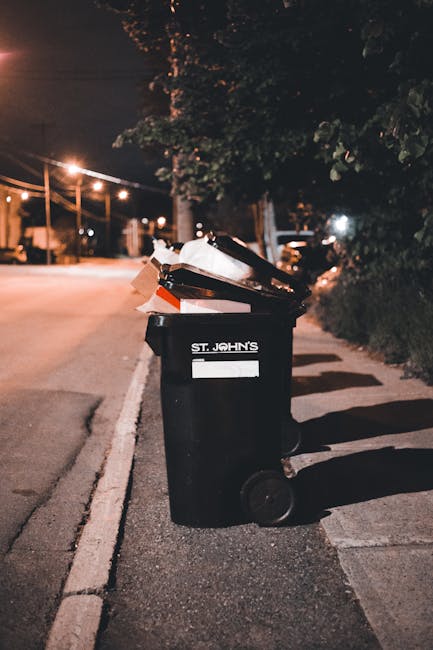 A black wheeled rubbish bin with the label 'St. John's' positioned on a paved sidewalk next to a curb in an outdoor urban setting at night. The bin is filled with various discarded waste materials, including cardboard boxes, paper, and plastic items, some of which are torn or crumpled. The background shows a street illuminated by streetlights, with a row of trees and bushes on the right side, and distant parked cars and faint building lights visible further down the road. The environment suggests a scene of residential or commercial waste collection, possibly indicating the use of local or private waste disposal services such as those provided by Rubbish Removal Swiss Cottage, rather than traditional municipal rubbish collection. The overall scene is quiet, with soft lighting emphasizing the details of the waste and the surrounding area, conveying the practical aspects of rubbish disposal in an urban environment in the evening.