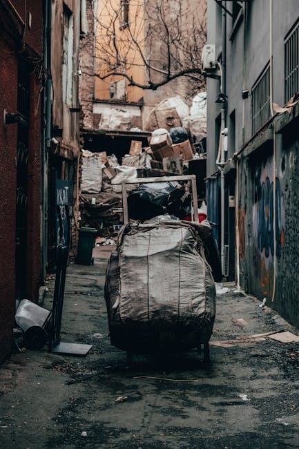 A narrow urban alleyway cluttered with overflowing piles of rubbish and discarded cardboard boxes at the back of buildings. In the foreground, a large, weathered, grey waste bag with visible creases and dirt stains is placed on a wheeled trolley, positioned centrally on a dark, grimy pavement. To the left, a small black trash bin lies on its side near the alley's edge, while to the right, parts of a grey metal fence and building wall are visible, with graffiti and vertical metal bars partially obscured by the clutter. In the background, stacked debris including cardboard boxes, black garbage bags, and mixed waste materials create a chaotic scene against the back wall of the alley, which is lined with bare, leafless trees and aged, faded brick buildings. The natural lighting appears dull and overcast, emphasizing the overall untidiness, suggesting an informal or private waste collection area that could potentially benefit from professional rubbish removal services such as those offered by Rubbish Removal Swiss Cottage to effectively manage urban waste and maintain cleanliness.