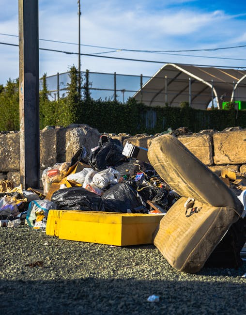 A black wheeled rubbish bin with the label 'St. John's' positioned on a paved sidewalk next to a curb in an outdoor urban setting at night. The bin is filled with various discarded waste materials, including cardboard boxes, paper, and plastic items, some of which are torn or crumpled. The background shows a street illuminated by streetlights, with a row of trees and bushes on the right side, and distant parked cars and faint building lights visible further down the road. The environment suggests a scene of residential or commercial waste collection, possibly indicating the use of local or private waste disposal services such as those provided by Rubbish Removal Swiss Cottage, rather than traditional municipal rubbish collection. The overall scene is quiet, with soft lighting emphasizing the details of the waste and the surrounding area, conveying the practical aspects of rubbish disposal in an urban environment in the evening.