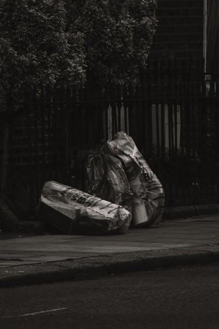 Two large black trash bags made of plastic, filled and tied at the top, are placed on the edge of a sidewalk in front of a black metal fence with vertical bars. The bags appear slightly crumpled and contain waste materials, suggesting they are for rubbish collection. Behind the fence, there are dense leafy bushes or trees, adding greenery to the scene, and a dark building or structure is partially visible in the background. The pavement shows slight wear, and the overall lighting indicates an overcast or shaded environment, typical of an urban setting near residential or commercial properties where private waste disposal and rubbish removal services, such as those offered by Rubbish Removal Swiss Cottage, might be used for local rubbish clearance.