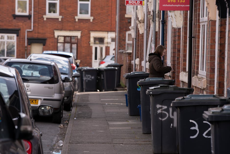 A residential street scene featuring a row of dark grey wheelie bins lined along the pavement outside brick houses, with a woman in a brown coat standing near one of the bins, appearing to be depositing or retrieving rubbish. Several cars are parked along the street, including a silver hatchback closest to the foreground, with others extending into the background. The houses have reddish-brown brick facades with white-framed windows and some decorative stonework around the windows and doorways. In the background, a red “SOLD” sign is visible, indicating the property has been sold. The overall scene depicts typical urban rubbish disposal arrangements, with the bins positioned for collection, representing a common aspect of private waste management or alternative rubbish collection services provided by companies like Rubbish Removal Swiss Cottage.